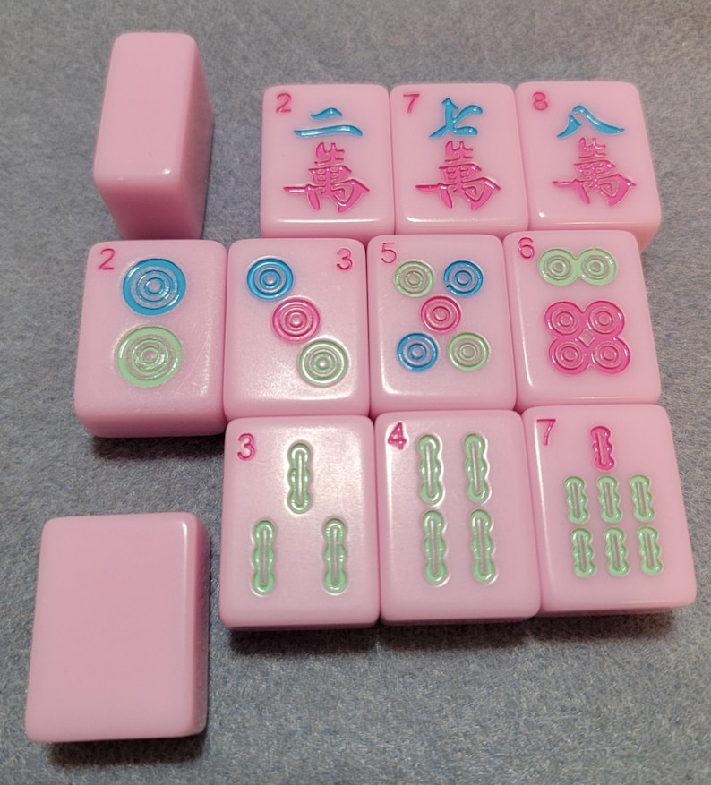 May include: A collection of pink Mahjong tiles with various symbols and numbers in blue, green, and pink. The tiles are arranged on a gray surface, showcasing the game's components.