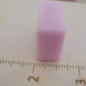 May include: A light purple rectangular object, possibly a bead or a small block, is positioned on a wooden surface. The object is smooth and solid, with rounded edges. A ruler is visible in the foreground, providing a sense of scale.