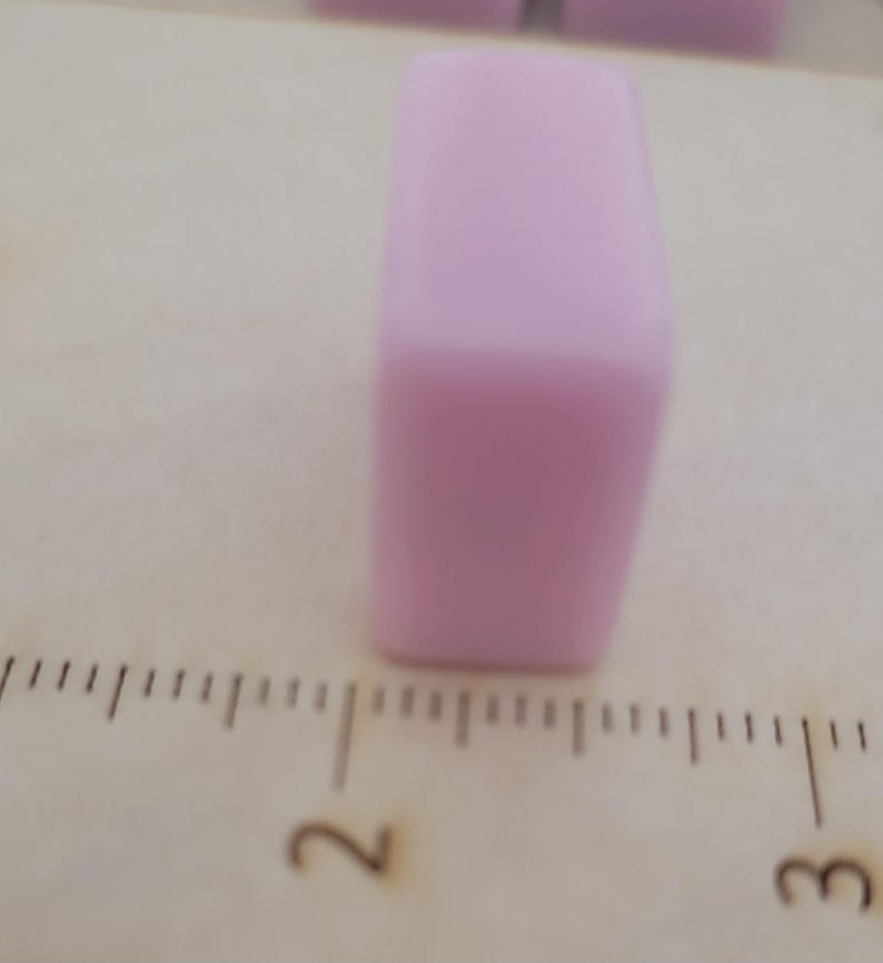 May include: A light purple rectangular object, possibly a bead or a small block, is positioned on a wooden surface. The object is smooth and solid, with rounded edges. A ruler is visible in the foreground, providing a sense of scale.