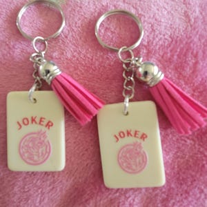 May include: Two keychains with rectangular cream-colored pendants that read "JOKER" in red, with a pink design. Each keychain has a pink tassel and a silver keyring.