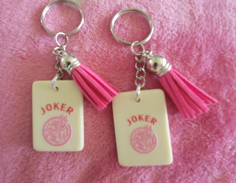 May include: Two keychains with rectangular cream-colored pendants that read "JOKER" in red, with a pink design. Each keychain has a pink tassel and a silver keyring.