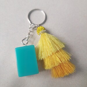 May include: A keychain with a rectangular, turquoise charm and a bright yellow tassel. The tassel is layered and attached to a silver-colored chain and keyring. The top of the tassel is embellished with small, clear rhinestones.