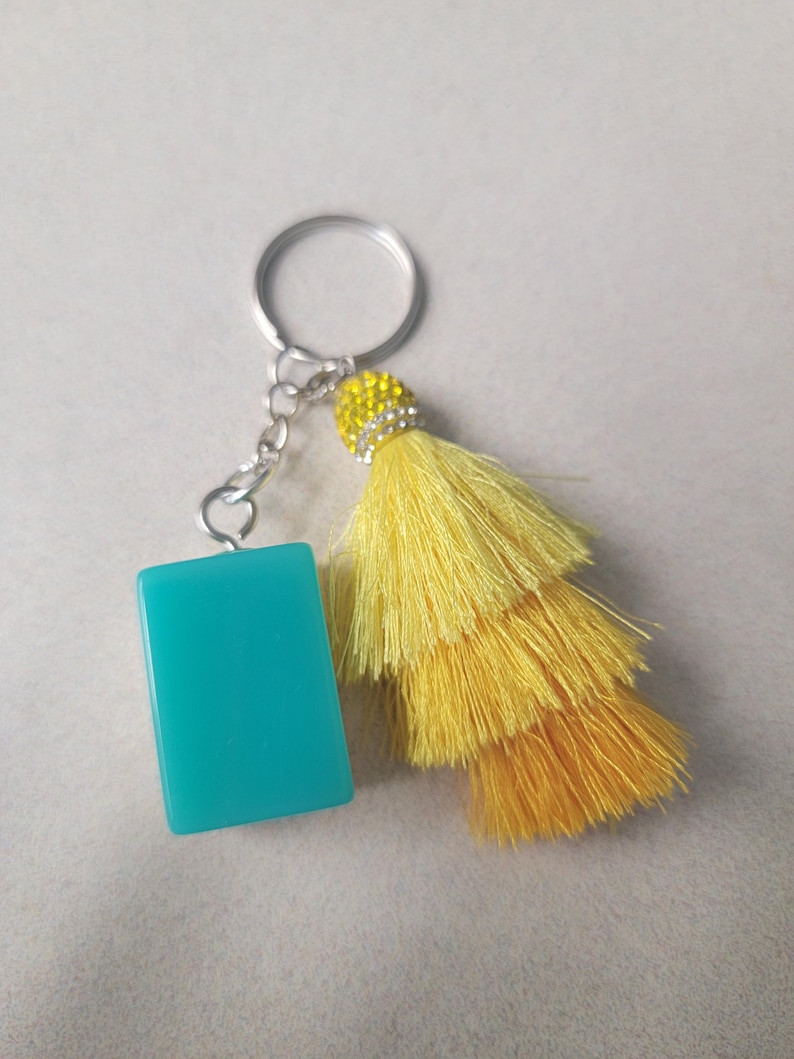 May include: A keychain with a rectangular, turquoise charm and a bright yellow tassel. The tassel is layered and attached to a silver-colored chain and keyring. The top of the tassel is embellished with small, clear rhinestones.