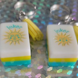 May include: Two keyrings with rectangular charms. Each charm has a white background with a yellow sun design and the word "Joker" in teal. The bottom of each charm is teal and yellow. Each keyring has a yellow tassel.
