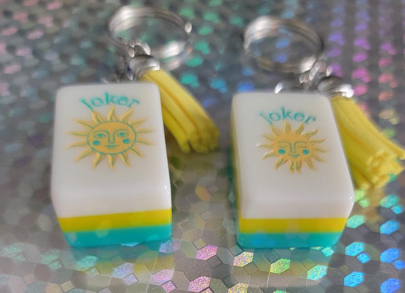 May include: Two keyrings with rectangular charms. Each charm has a white background with a yellow sun design and the word "Joker" in teal. The bottom of each charm is teal and yellow. Each keyring has a yellow tassel.