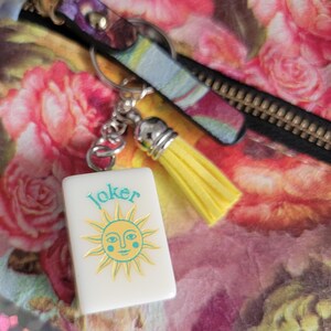 May include: A white rectangular keyring with the word "Joker" and a sun design in yellow and teal. The keyring is attached to a silver chain with a yellow tassel. The background features a floral pattern.