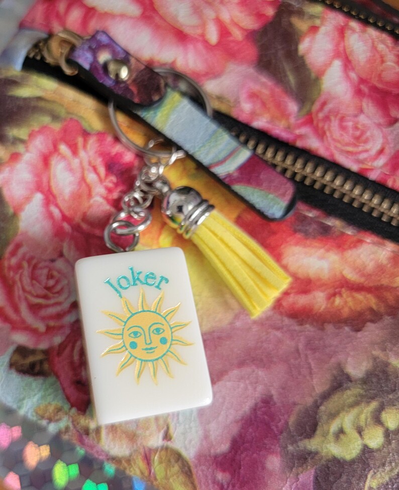 May include: A white rectangular keyring with the word "Joker" and a sun design in yellow and teal. The keyring is attached to a silver chain with a yellow tassel. The background features a floral pattern.