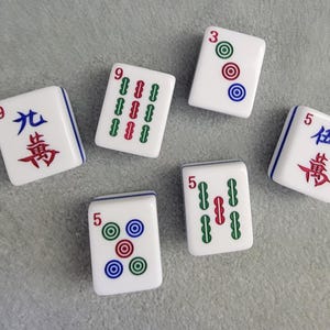 May include: Six white Mahjong tiles with blue borders are arranged on a gray surface. The tiles display various combinations of numbers, Chinese characters, and colored circles and lines. The tiles are for the classic game of Mahjong.