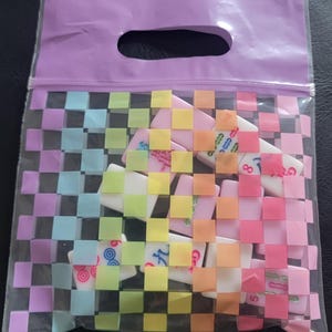 May include: A clear plastic bag with a woven rainbow pattern, containing several dominoes. The bag has a purple handle and zipper closure. The dominoes are white with colorful markings.