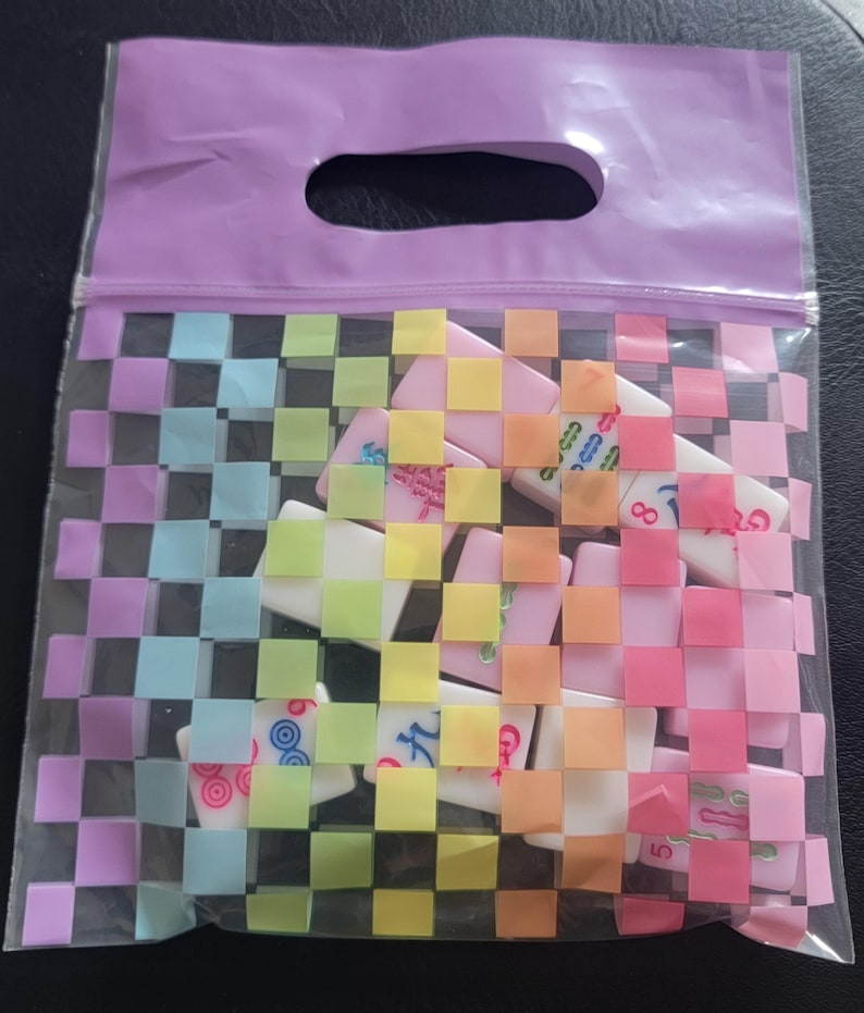 May include: A clear plastic bag with a woven rainbow pattern, containing several dominoes. The bag has a purple handle and zipper closure. The dominoes are white with colorful markings.