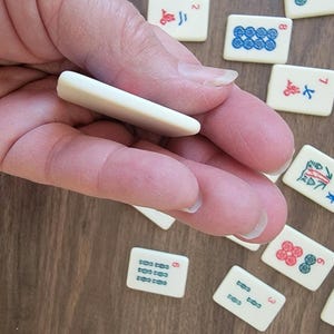 May include: A hand holding a white tile, a common component of a Mahjong set. The tile is blank and has no markings. The hand is resting on a wooden surface with other Mahjong tiles scattered around.