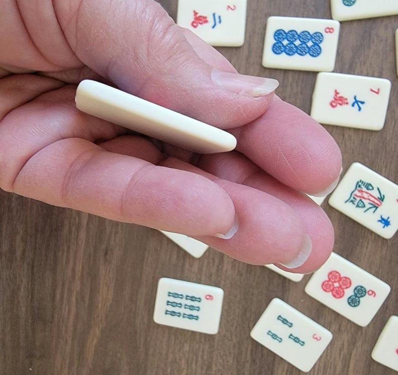 May include: A hand holding a white tile, a common component of a Mahjong set. The tile is blank and has no markings. The hand is resting on a wooden surface with other Mahjong tiles scattered around.