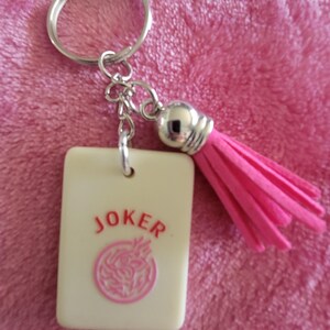 May include: A keychain featuring a rectangular cream-colored charm with the word "JOKER" in red and a pink circular design. The keychain includes a silver-colored ring, chain, and a pink tassel.