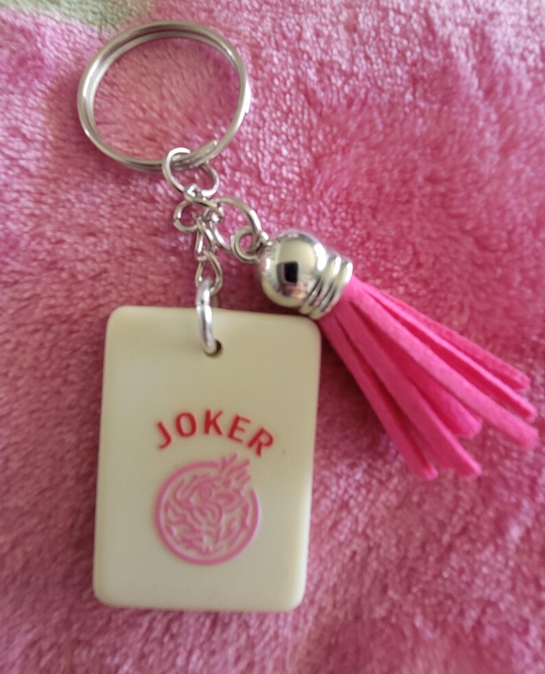 May include: A keychain featuring a rectangular cream-colored charm with the word "JOKER" in red and a pink circular design. The keychain includes a silver-colored ring, chain, and a pink tassel.