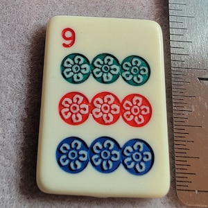 May include: A white domino tile with the number 9 printed in red. The tile has three rows of three circles each, with a white flower design inside each circle. The top row of circles is green, the middle row is red, and the bottom row is blue.