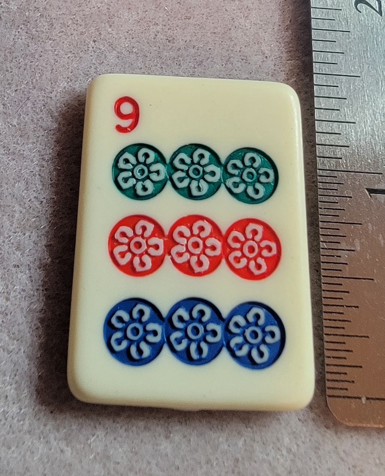 May include: A white domino tile with the number 9 printed in red. The tile has three rows of three circles each, with a white flower design inside each circle. The top row of circles is green, the middle row is red, and the bottom row is blue.