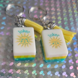 May include: Two keyrings with white rectangular charms featuring a sun design and the word "Joker". The charms have yellow, blue, and green accents. Each keyring has a silver ring and a yellow tassel.