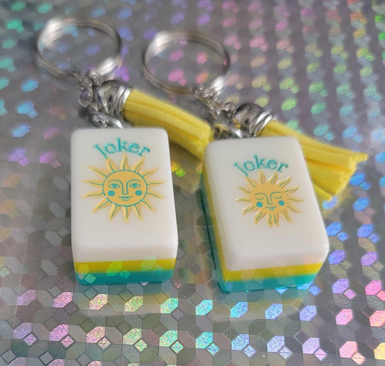May include: Two keyrings with white rectangular charms featuring a sun design and the word "Joker". The charms have yellow, blue, and green accents. Each keyring has a silver ring and a yellow tassel.