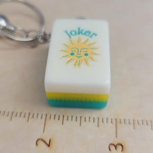 May include: A white rectangular keychain charm with the word "Joker" in teal above a smiling sun design. The bottom edge has a yellow and teal stripe. A silver-coloured keyring is attached.