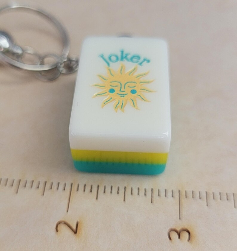 May include: A white rectangular keychain charm with the word "Joker" in teal above a smiling sun design. The bottom edge has a yellow and teal stripe. A silver-coloured keyring is attached.