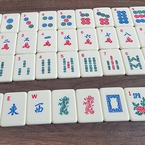 May include: A set of 36 white mahjong tiles with blue and red Chinese characters and symbols. The tiles are arranged in rows and columns on a wooden surface. The tiles are numbered 1 through 9 and include wind, dragon, and flower tiles.