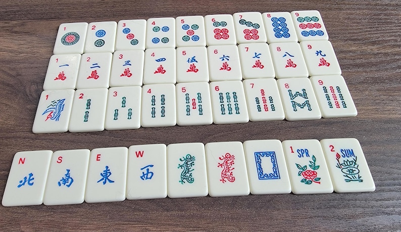 May include: A set of 36 white mahjong tiles with blue and red Chinese characters and symbols. The tiles are arranged in rows and columns on a wooden surface. The tiles are numbered 1 through 9 and include wind, dragon, and flower tiles.