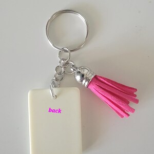 May include: A keychain with a rectangular, off-white tag that reads "back" in pink lettering. The keychain has a silver-colored ring and chain, and a bright pink tassel. The tassel is made of a soft, fringed material.