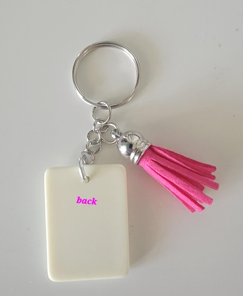 May include: A keychain with a rectangular, off-white tag that reads "back" in pink lettering. The keychain has a silver-colored ring and chain, and a bright pink tassel. The tassel is made of a soft, fringed material.