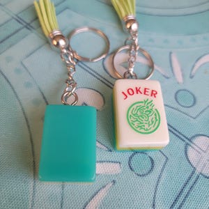 May include: Two keychains with silver-tone hardware and light green tassels. One keychain has a teal rectangular charm, the other has a white tile with the word "JOKER" and a green design. The keychains are on a patterned blue surface.