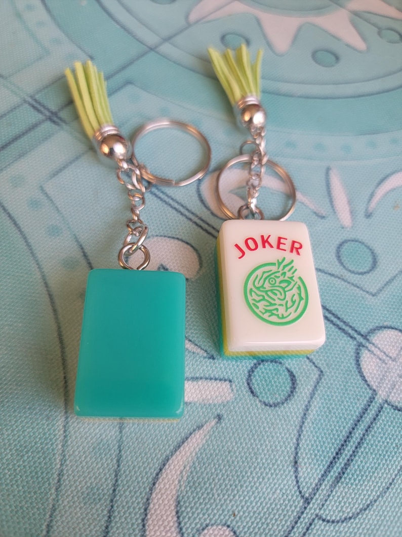 May include: Two keychains with silver-tone hardware and light green tassels. One keychain has a teal rectangular charm, the other has a white tile with the word "JOKER" and a green design. The keychains are on a patterned blue surface.