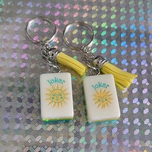 May include: Two keyrings with white rectangular charms featuring a sun design and the word "Joker" in teal. The charms have a yellow border and are attached to silver keyrings with yellow tassels.