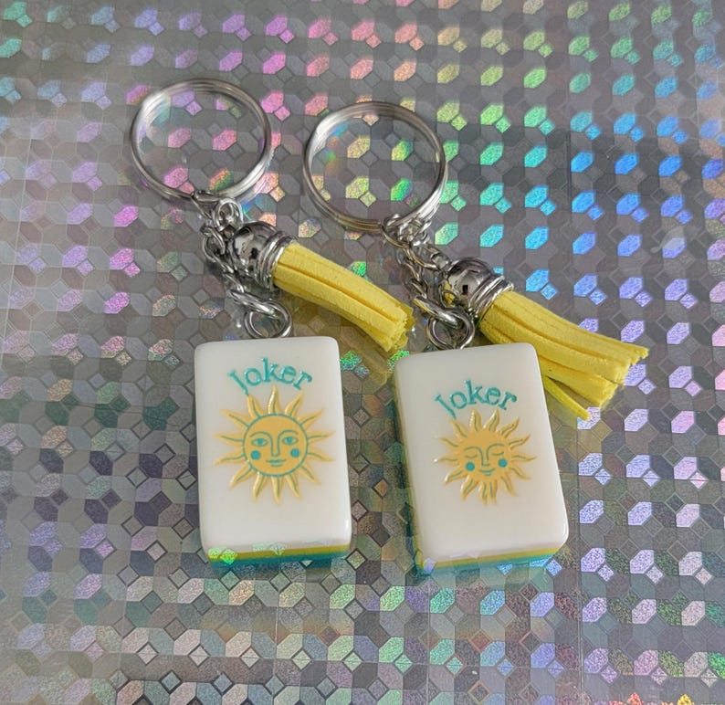 May include: Two keyrings with white rectangular charms featuring a sun design and the word "Joker" in teal. The charms have a yellow border and are attached to silver keyrings with yellow tassels.