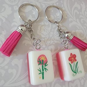 May include: Two keychains with silver rings, pink tassels, and square charms. The charms have a white background with a pink border and feature floral designs. One has a bamboo-like design, and the other has a red flower.
