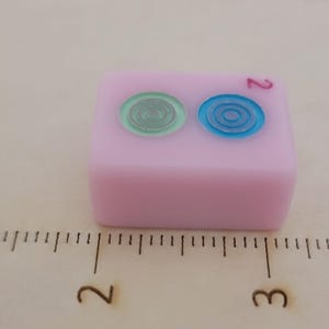 May include: A pink Mahjong tile with two circular designs in green and blue. The number 2 is printed in red. The tile is on a wooden surface with a ruler.