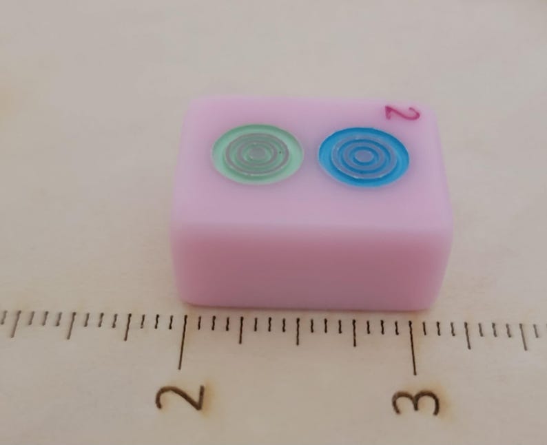 May include: A pink Mahjong tile with two circular designs in green and blue. The number 2 is printed in red. The tile is on a wooden surface with a ruler.