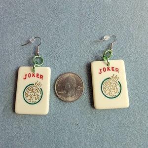 May include: A pair of white, square-shaped earrings with a green and gold design. The earrings feature the word "JOKER" in red lettering and a green circle with a gold design inside. The earrings are attached to silver hooks with green rings.