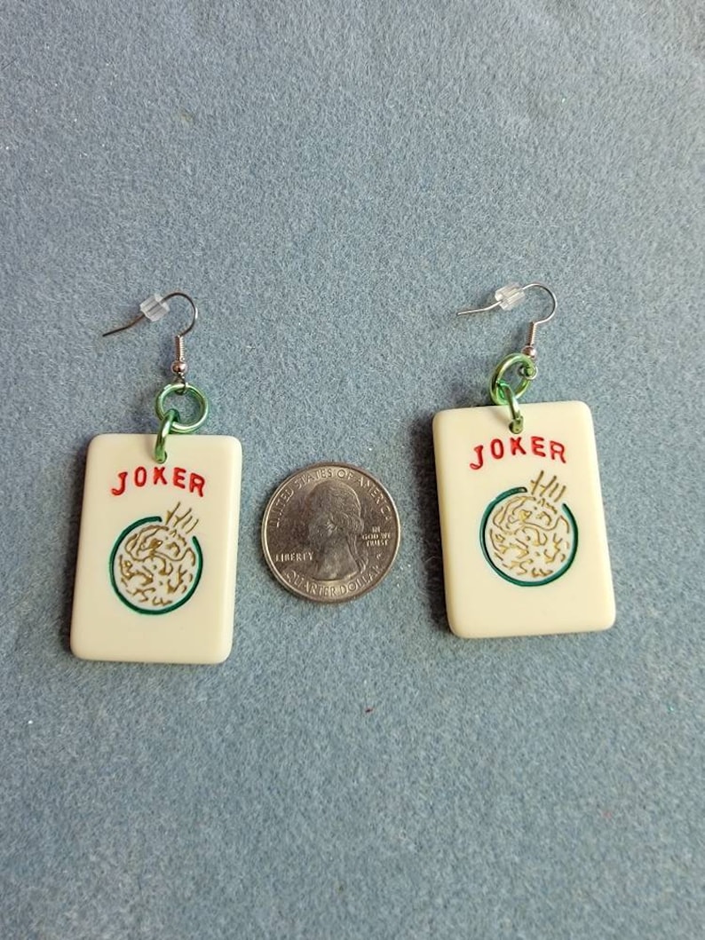 May include: A pair of white, square-shaped earrings with a green and gold design. The earrings feature the word "JOKER" in red lettering and a green circle with a gold design inside. The earrings are attached to silver hooks with green rings.