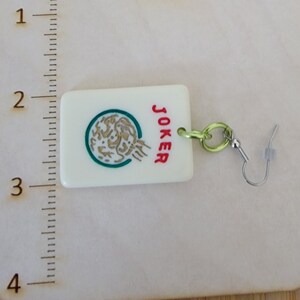 May include: White ceramic earring with a green and gold design and the word "JOKER" in red. The earring is attached to a silver hook.