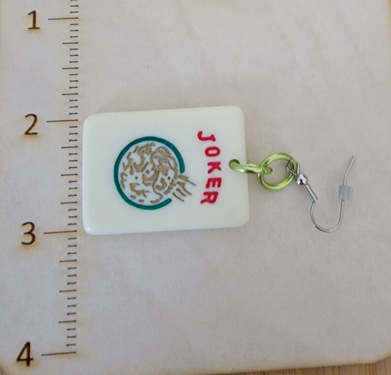 May include: White ceramic earring with a green and gold design and the word "JOKER" in red. The earring is attached to a silver hook.