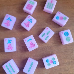 May include: A collection of pink Mahjong tiles with various symbols and numbers in green, blue, and pink. The tiles are scattered on a wooden surface, showcasing the game's components.