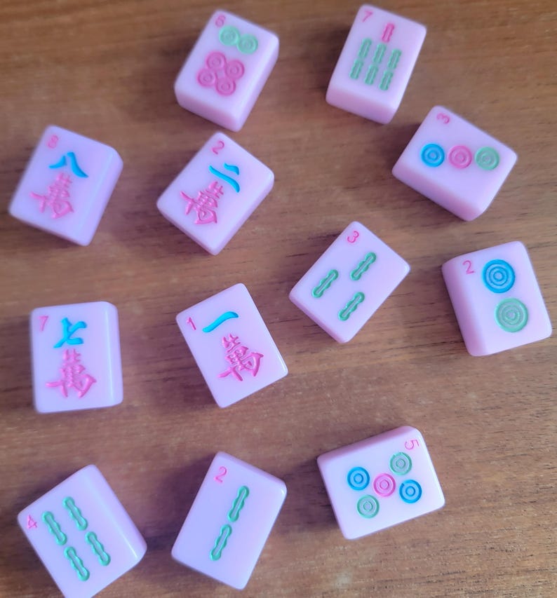 May include: A collection of pink Mahjong tiles with various symbols and numbers in green, blue, and pink. The tiles are scattered on a wooden surface, showcasing the game's components.