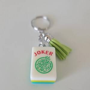 May include: A white Mahjong tile keychain with the word "JOKER" in red and a green dragon design. The tile has a green, yellow, and blue border, and a light green tassel. The keychain is attached to a silver keyring.