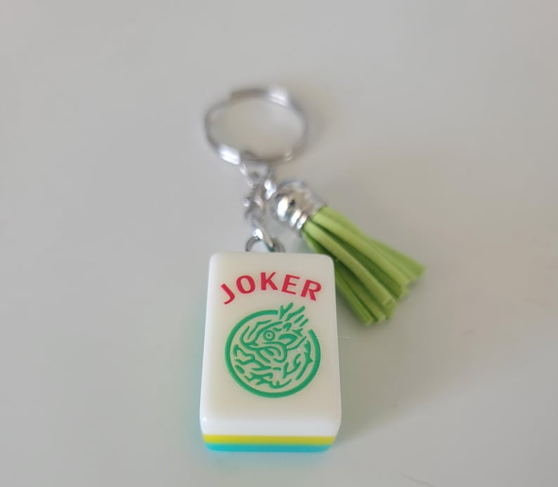 May include: A white Mahjong tile keychain with the word "JOKER" in red and a green dragon design. The tile has a green, yellow, and blue border, and a light green tassel. The keychain is attached to a silver keyring.