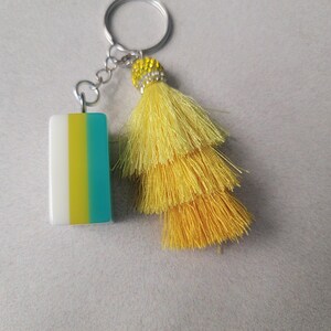 May include: A keychain with a rectangular charm and a yellow tassel. The charm is white, yellow, and teal. The tassel is made of layered yellow threads and has a decorative beaded cap. The keychain ring is silver.