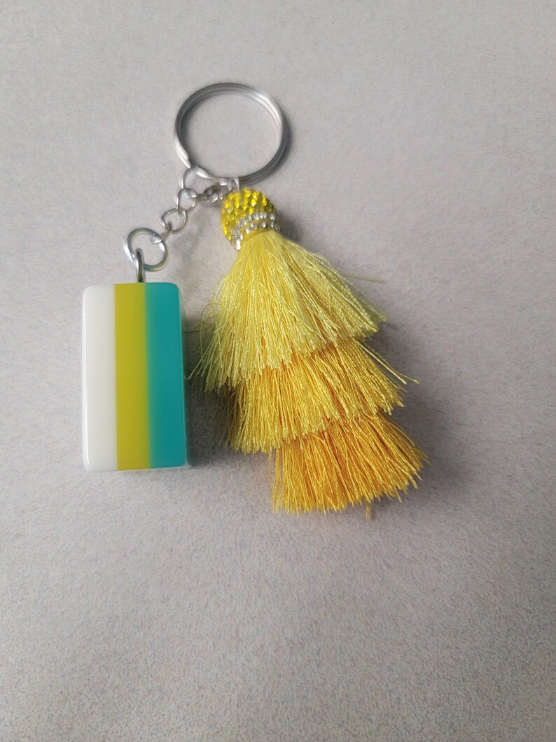 May include: A keychain with a rectangular charm and a yellow tassel. The charm is white, yellow, and teal. The tassel is made of layered yellow threads and has a decorative beaded cap. The keychain ring is silver.