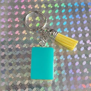 May include: A teal rectangular keyring with a silver ring and yellow tassel. The tassel has a silver cap. The keyring is a solid teal colour and is rectangular in shape.