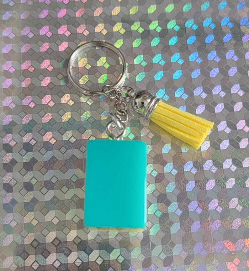 May include: A teal rectangular keyring with a silver ring and yellow tassel. The tassel has a silver cap. The keyring is a solid teal colour and is rectangular in shape.