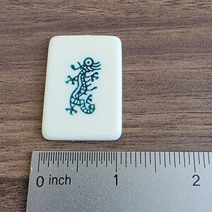 以下が含まれることがあります： 緑色のトカゲのデザインが施された白いタイル。トカゲは右を向いており、長い尻尾があります。