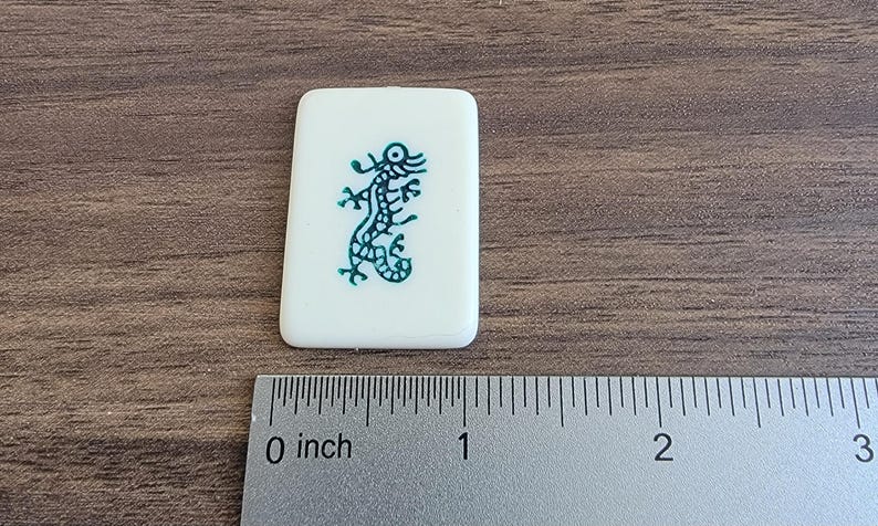 以下が含まれることがあります： 緑色のトカゲのデザインが施された白いタイル。トカゲは右を向いており、長い尻尾があります。