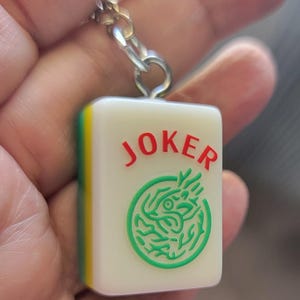 May include: A white Mahjong tile keychain with the word "JOKER" in red and a green dragon design. The tile has a yellow and green edge and is attached to a silver chain.